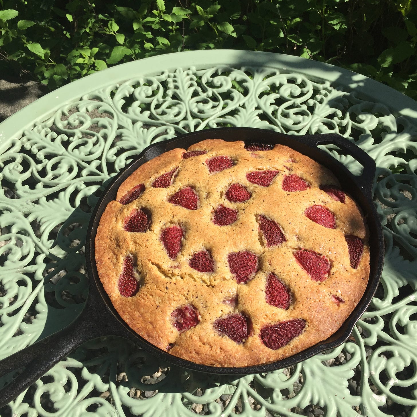On a mint green outdoor table of wrought iron, a cast iron pan full of a golden-brown cake flecked with strawberry halves rests in the sun.