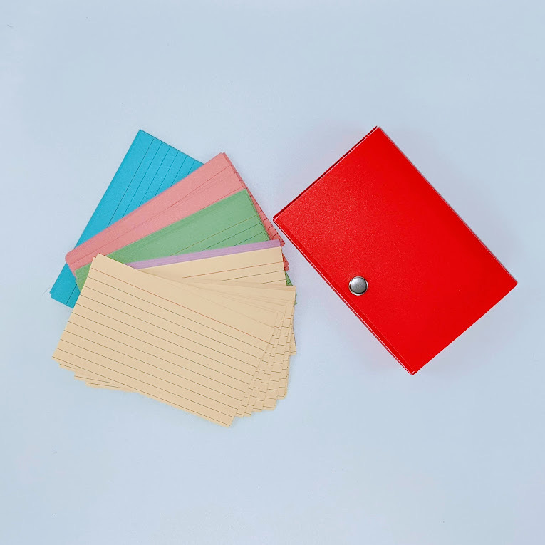 An image of a red plastic case for holding index cards and a stack of multicolored index cards (blue, pink, green, purple, and yellow).