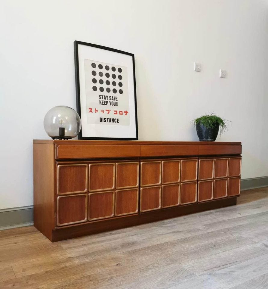 A stunning mid-century sideboard in brown wood with a grid-based system of decorative wooden accents on the doors of the cabinets. On top of the sideboard there's a vintage-style posted, a potted plant, and a space-age lamp. 
