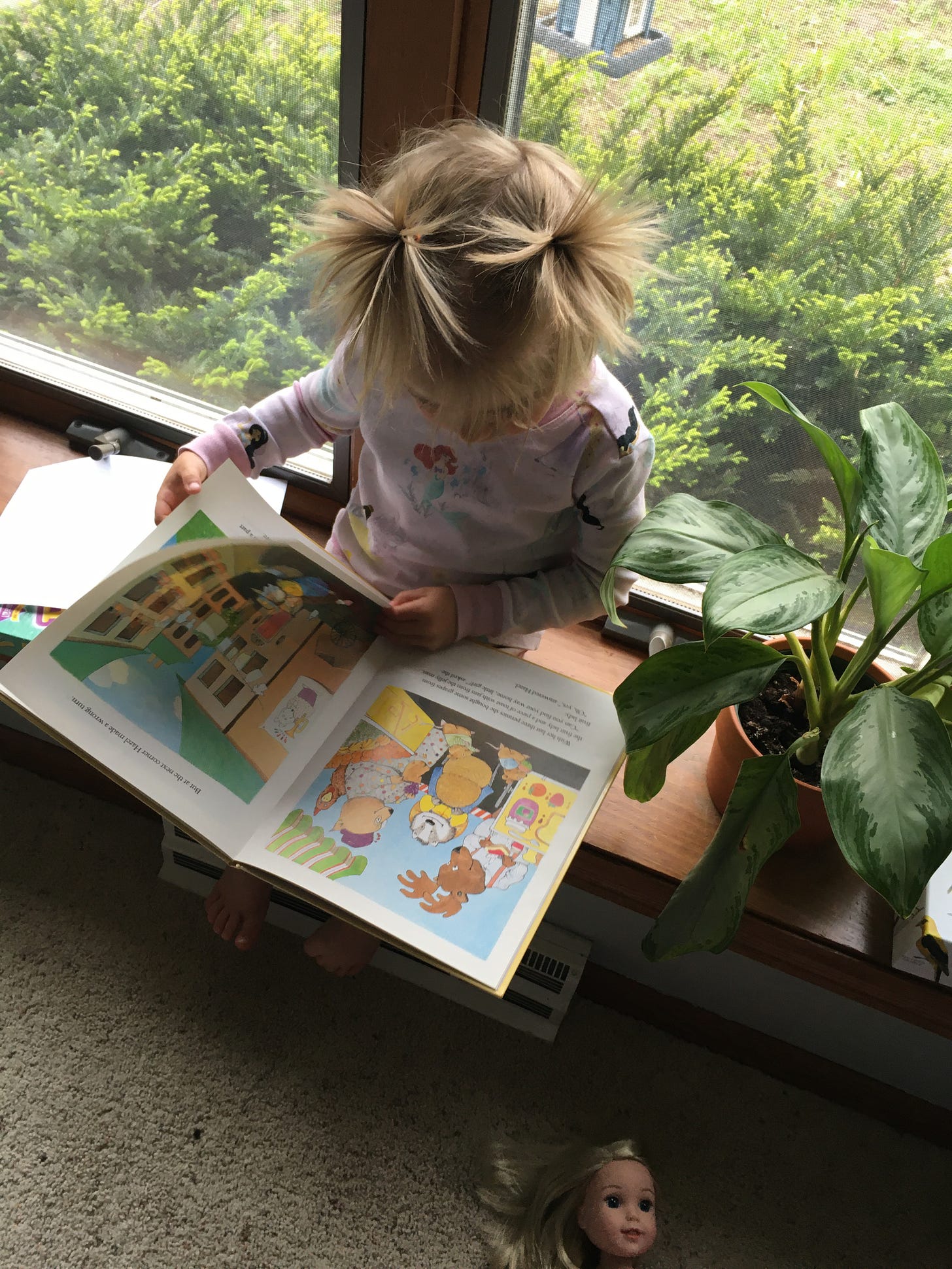A little girl sitting on a windowsill reading a book next to a plant