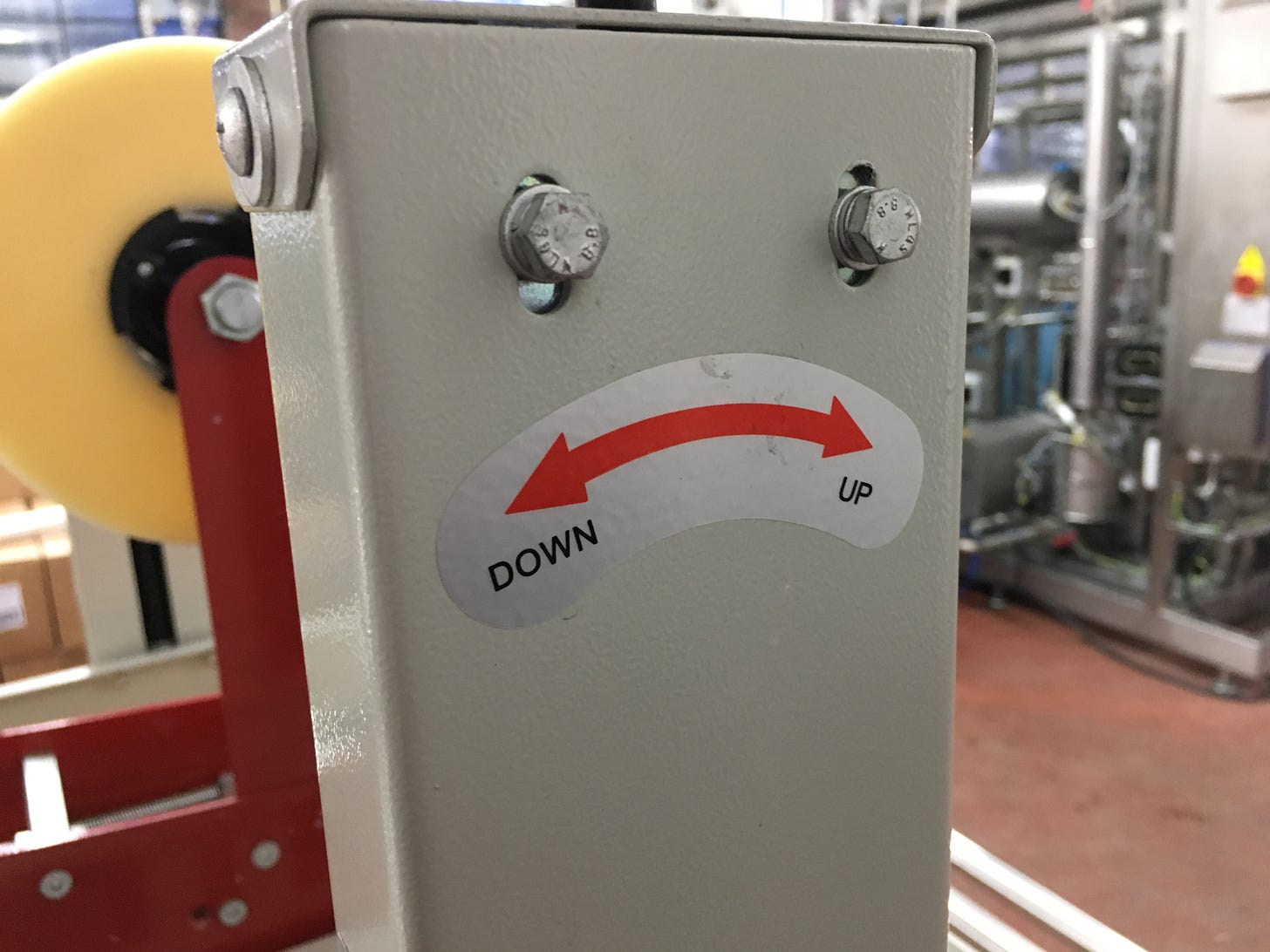 Side of a tape machine in a brewery packing facility, shows a red arrow in a downward arc with 'up' and 'down' written on either side. Two bolts above the arc create a sad face.