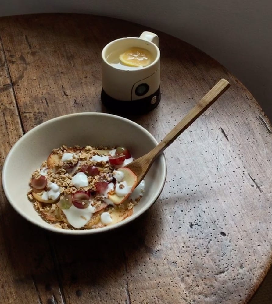 Set against the top of an antique dark wooden table: a bowl of cereal with grapes cut over it and yogurt blobs dotted. In it is a wooden spoon. Behind, up to the right, a mug with a slice of lemon submerged in the liquid. 