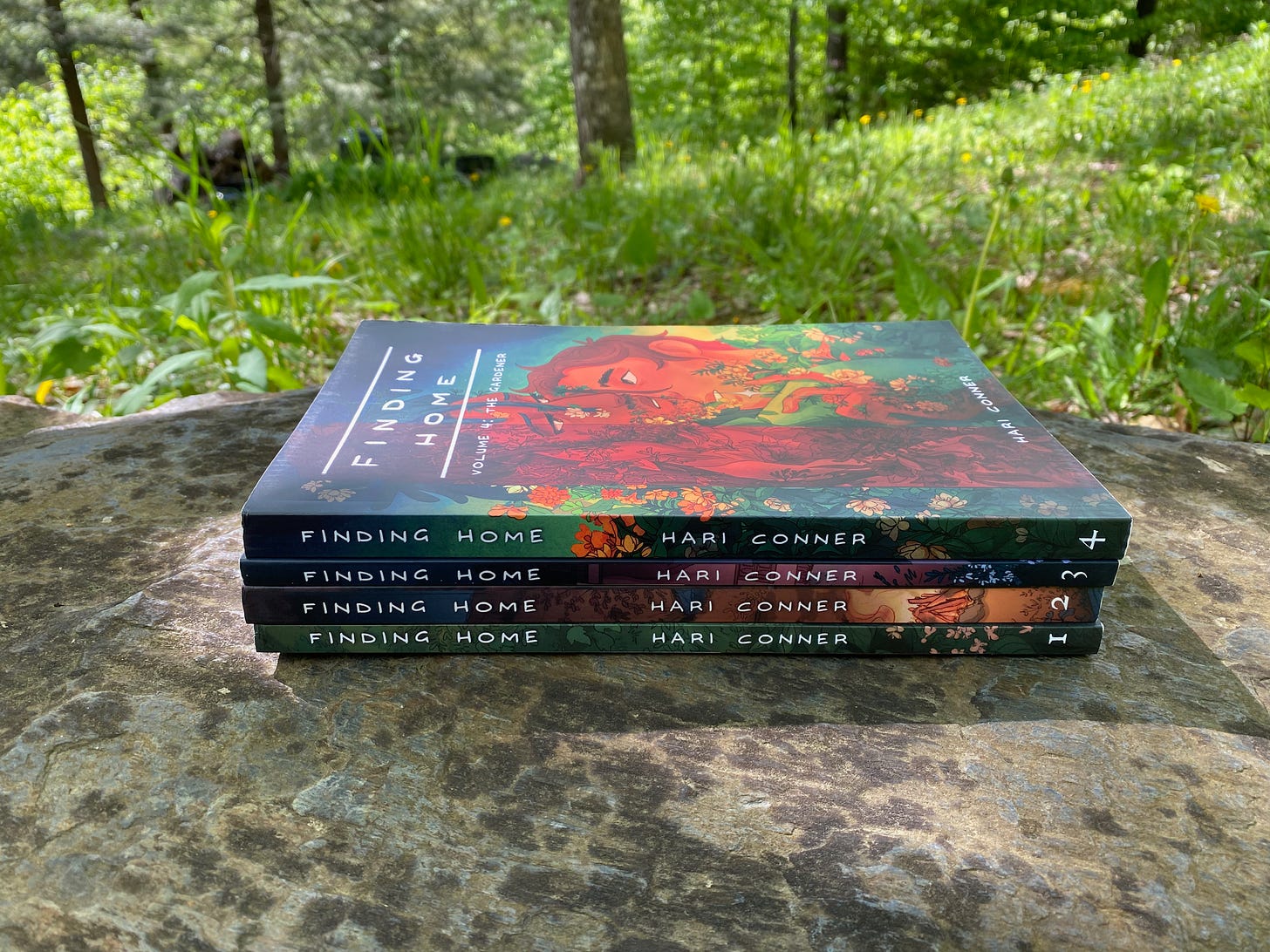 Four paperback volumes of Finding Home stacked on a flat rock, with bright grass and trees in the background.