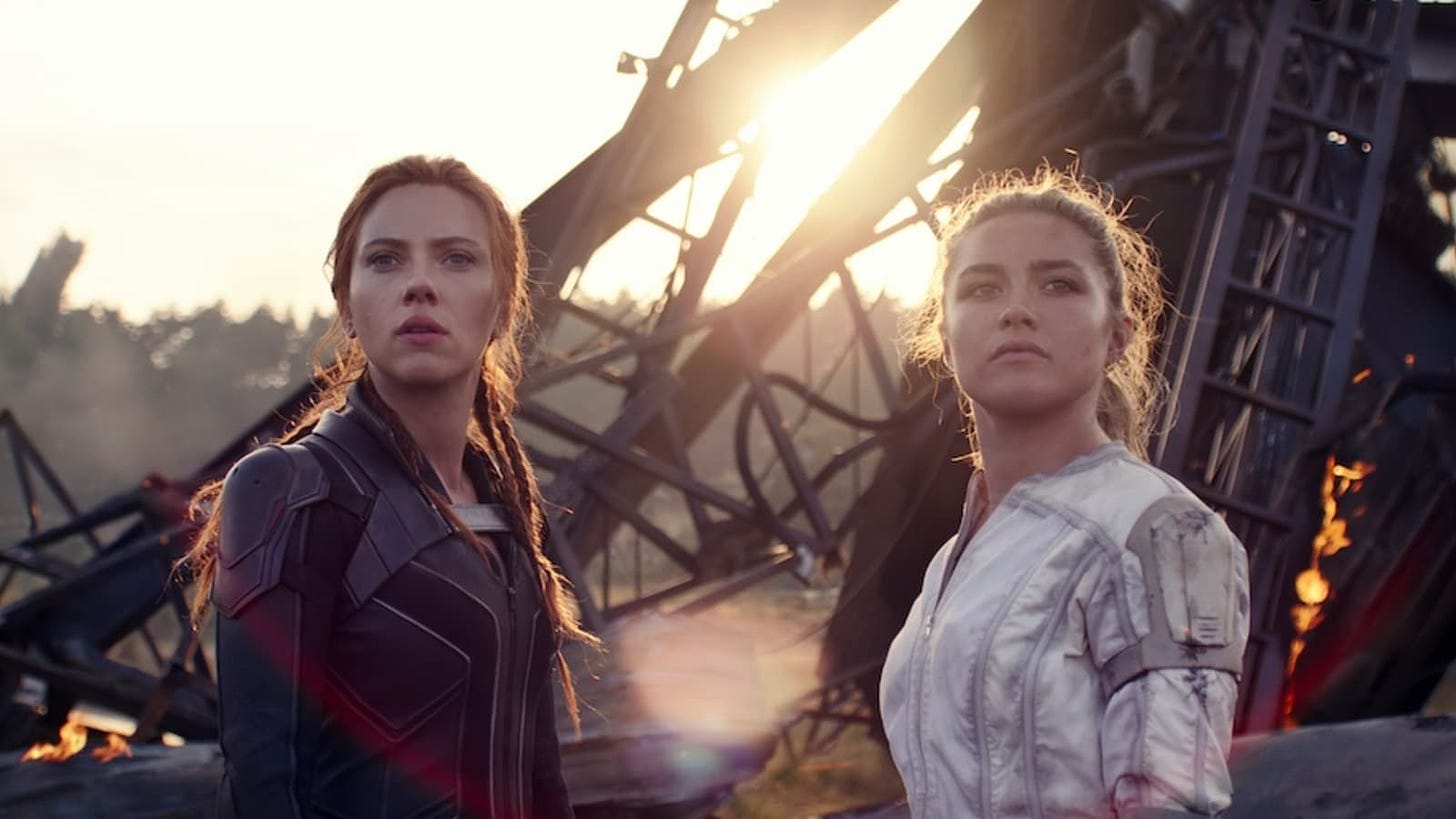 Black Widow and her sister stand and look at the camera with the wreckage of a plane behind them.