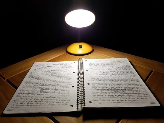 Foto colorida de um caderno grande aberto com as duas páginas escritas numa grafia fina em tinta preta. O caderno está aberto sobre uma mesa de madeira com um abajur aceso por perto. Todo o fundo da imagem está em completa escuridão.