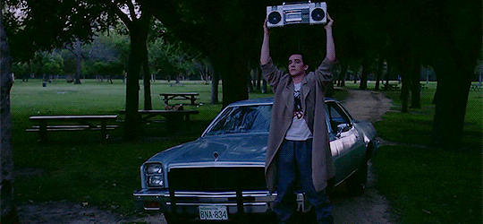 ID: Lloyd Dobler holds up a boombox in front of his car in an evergreen yard.