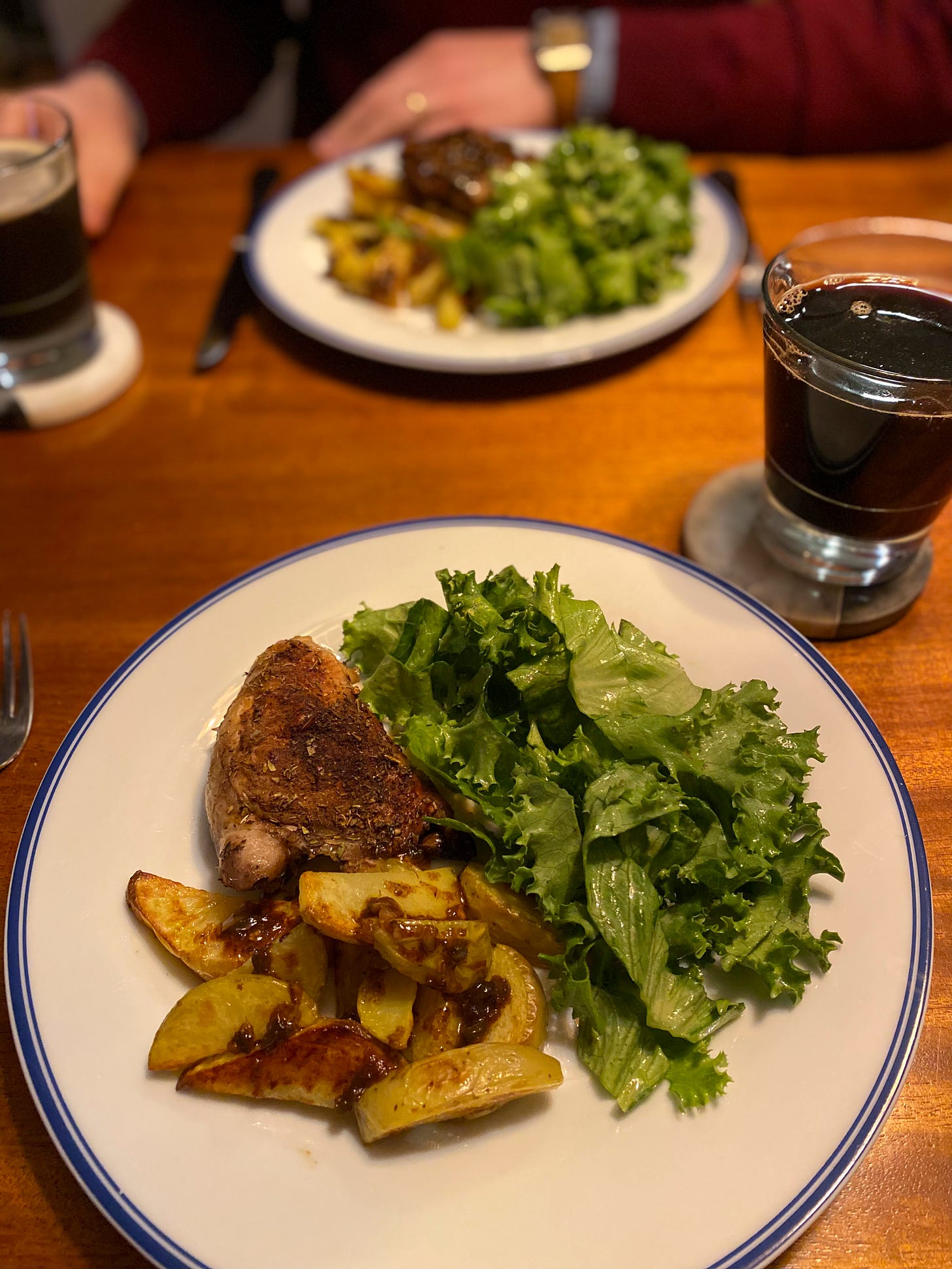 Two dinner plates across from each other, each filled with green salad, potato wedges with sauce drizzled on top, and a jerk chicken thigh with crispy-looking skin. Two glasses of dark beer rest on coasters near the tops of the plates.