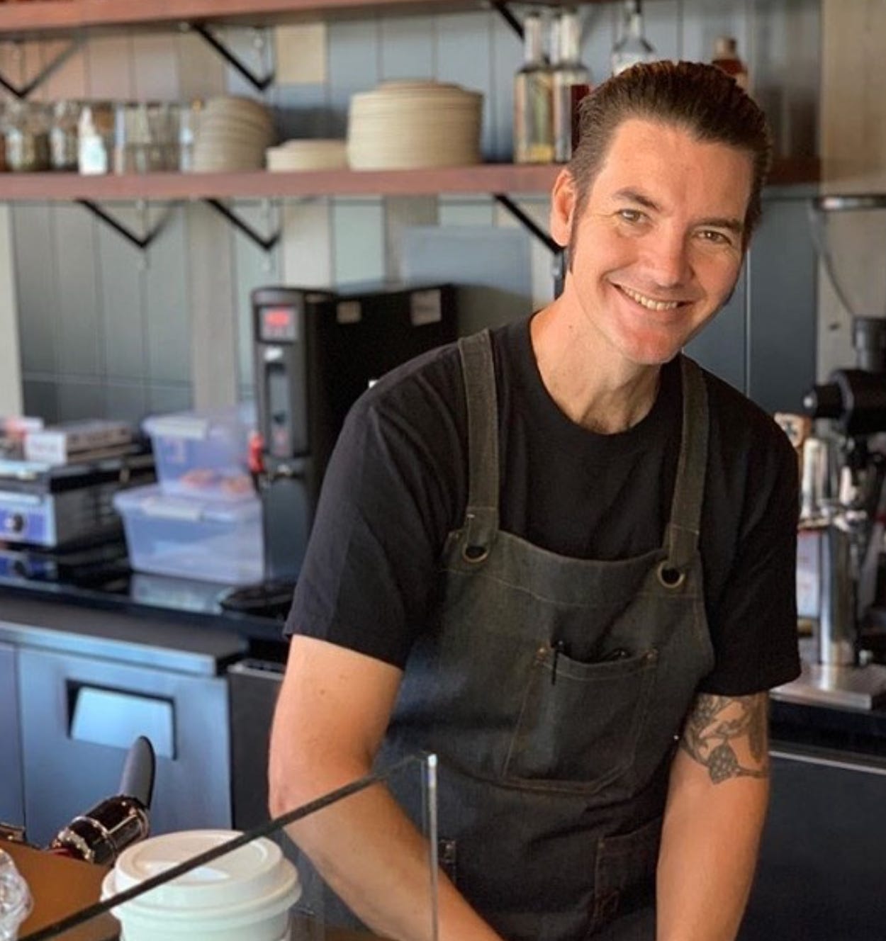 A tall, white barista with black hair slicked back wearing a white shirt and denim apron making a coffee drink and smiling at the camera.