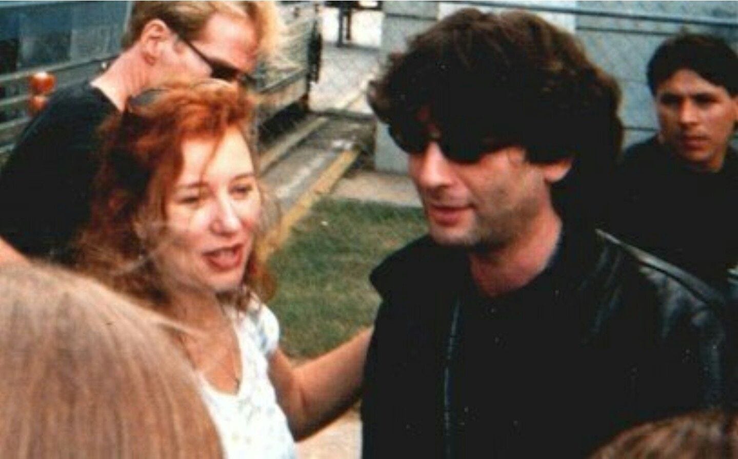 Foto antiga de Tori Amos, à esquerda, ruiva com um óculos sobre a cabeça, sorrindo e tocando as costas de Gaiman, à direita, cabelo preto bagunçado, óculos escuros e jaqueta preta. eles estão cercados de pessoas e ambos sorriem