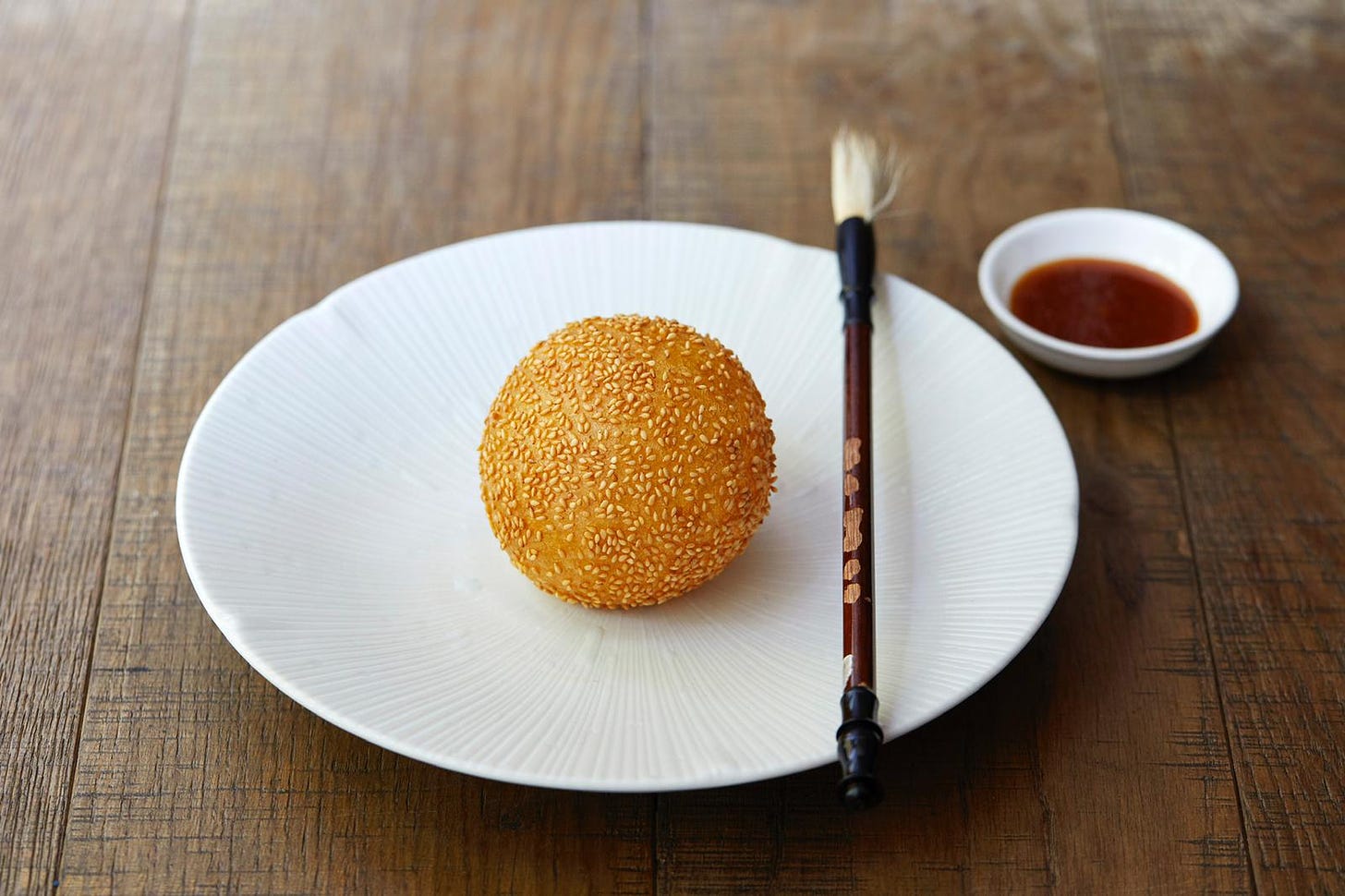 A puff ball pastry covered in sesame seeds on a small white plate. On the side of the plate is a paint brush and next to the plate is a small bowl of red coloured sauce. Plate, bowl and brush are on a wooden table