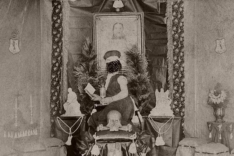 black and white photo showing a scribbled out figure sat on an elaborate and ornate chair with lots of decorations around them, including a portrait behind them and a skull in front of them