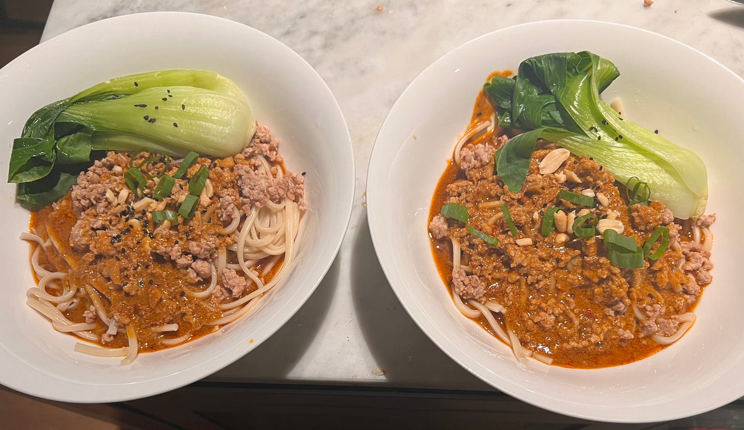 Two bowls of dan dan noodles, white udon noodles covered in reddish brown pork, each bowl has a green piece of bok choi.