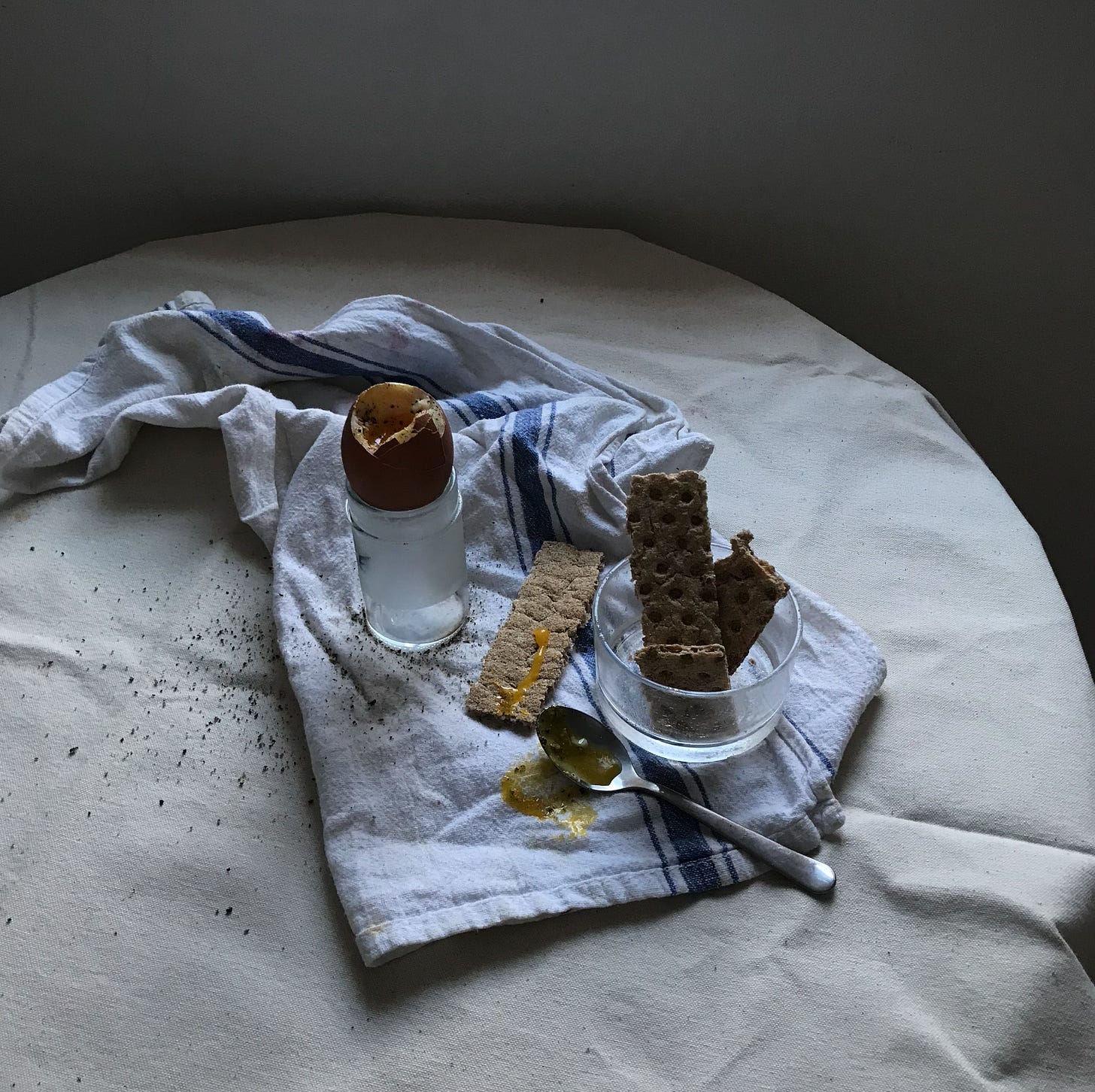 Atop a splayed white and blue tea towel, on a table covered in cream canvas, in evening light: a cracked boiled egg in a narrow glass, crispbreads stood up in a glass ramekin, an eggy metal spoon, pepper ground all over. 