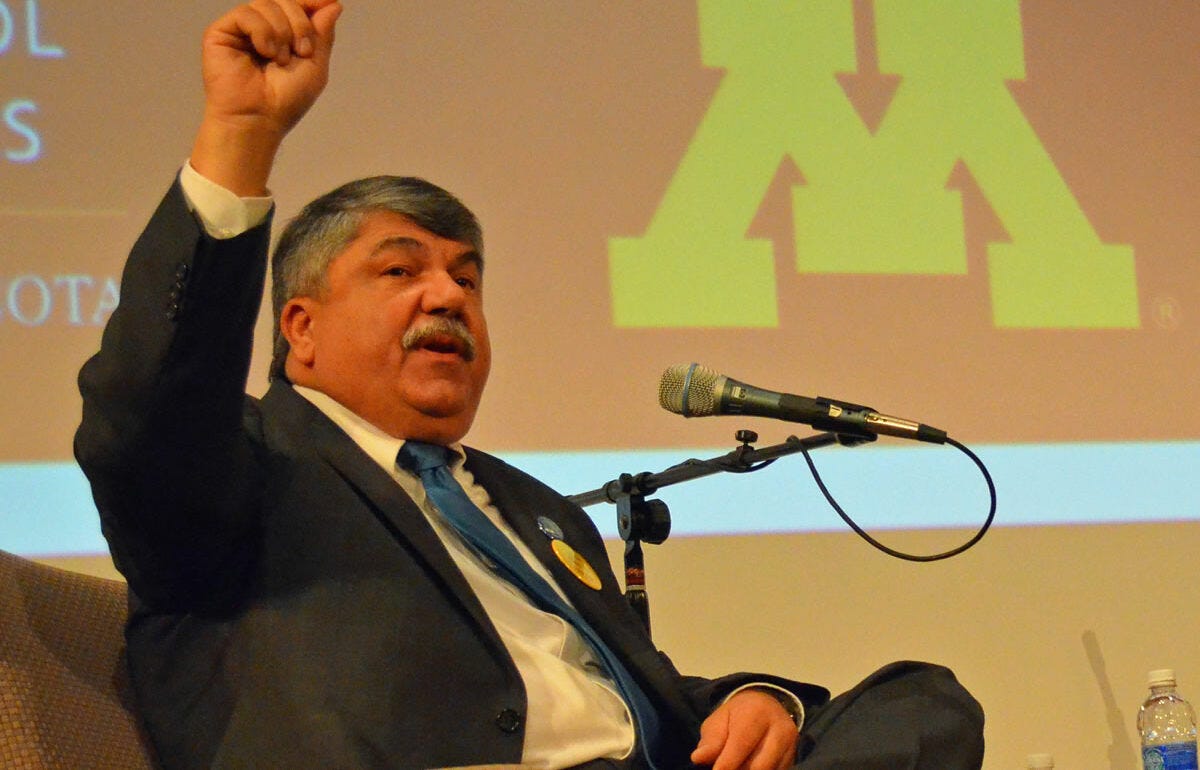 Richard Trumka sits in front of a microphone in a gray suit and blue tie gesturing to the ceiling with his hands, in front of a presentation with the U of M yellow "M" logo