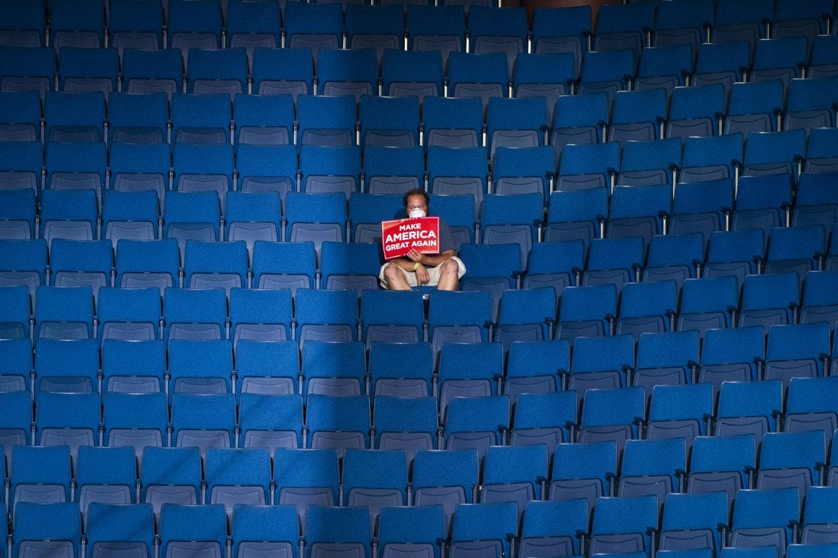 A lone Trump rally attendee sits alone with a bright red Trump sign, surrounded by empty blue stadium seats. 