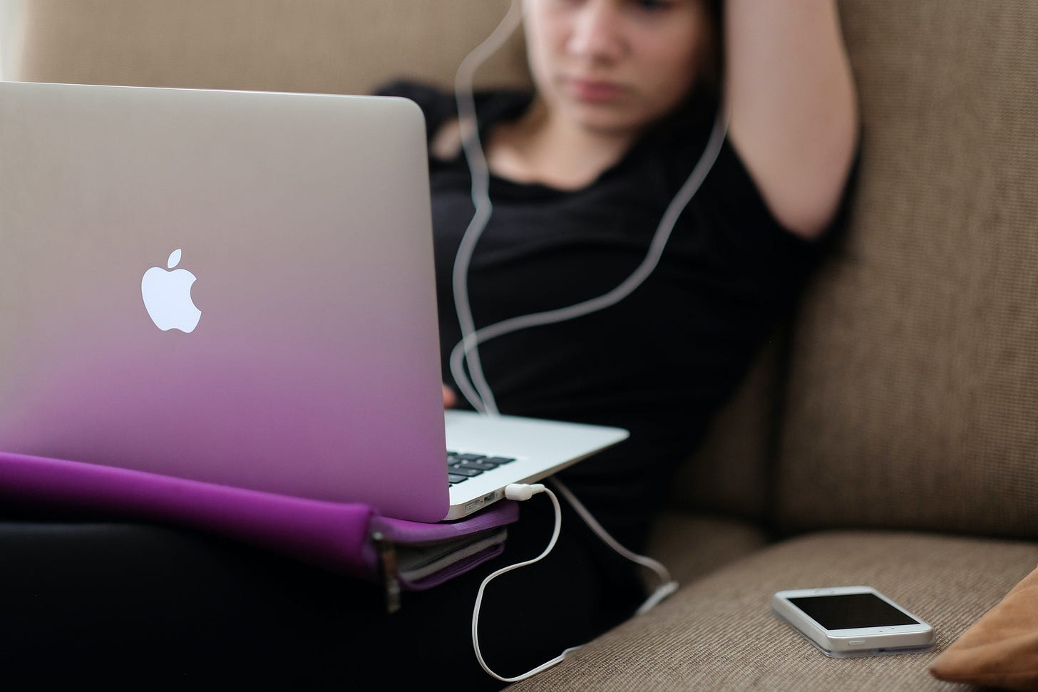 A young woman souches on a couch with a laptop on her lap, headphones in, her phone next to her.