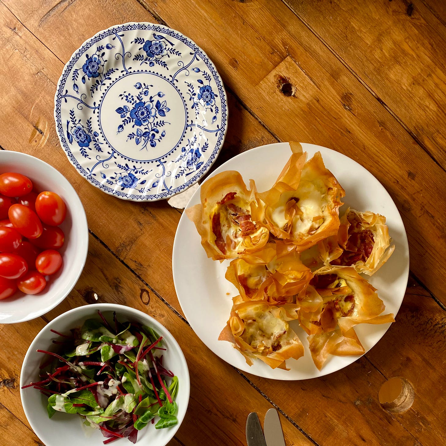 White plate holds six small filo tarts, next to a bowl of mixed salad with a separate bowl of cherry plum tomatoes and a small blue and white plate to the side
