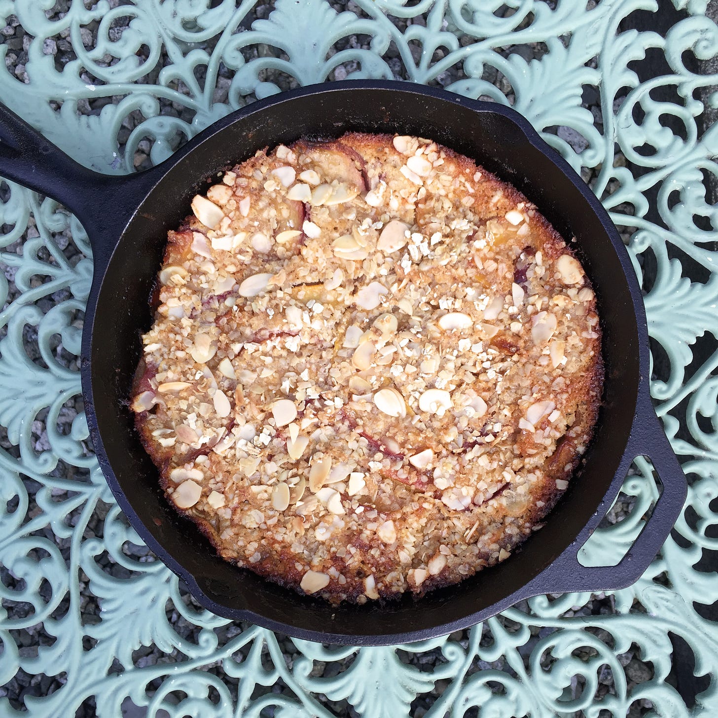 from above, a cast iron pan of peach cobbler topped with almonds and oats sits on a mint green wrought-iron table.