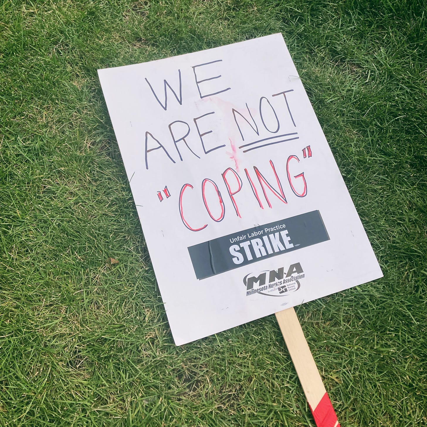 a picket sign lays on the grass and it reads "WE ARE NOT "COPING" in written marker. it also says "Unfair labor practice STRIKE" MNA Minnesota Nurses Assocation