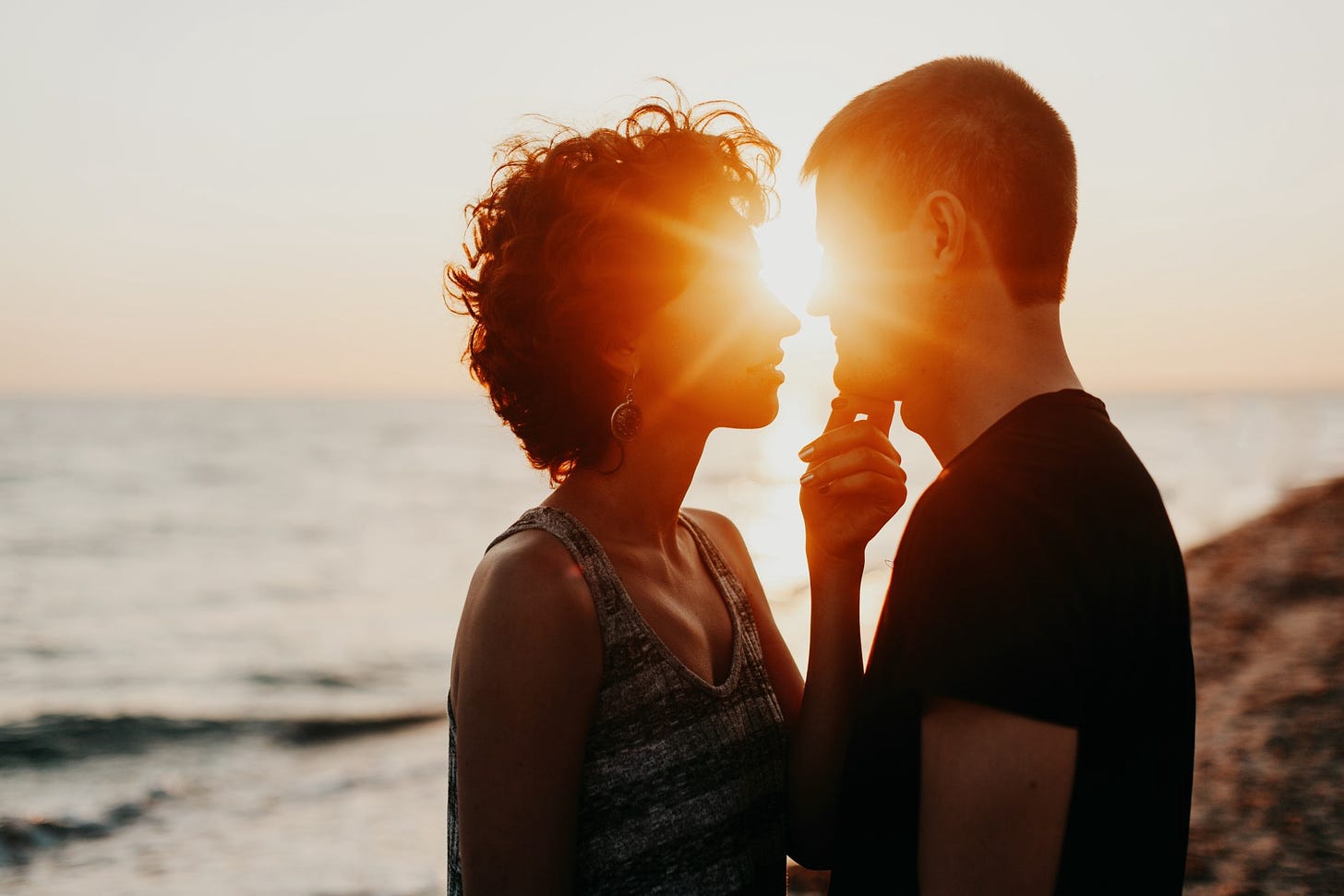 The silhoutte of a couple embraces on a beach as the sun sets in the background between them.