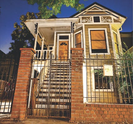 A two-story house stands behind an iron gate with red brick columns. The house is turn-of-the-century, featuring a long staircase which leads up to the front door. The sky is dark, indicating it is dusk when the picture was taken. 