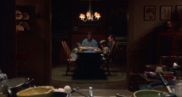 We are looking at the Hess family from outside a rectangular doorway as they sit, looking sad, around the dining table.