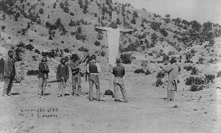 Penitente "crucifying" one of their own: person on huge wooden cross, which is held up by ropes, with men watching, 1888.