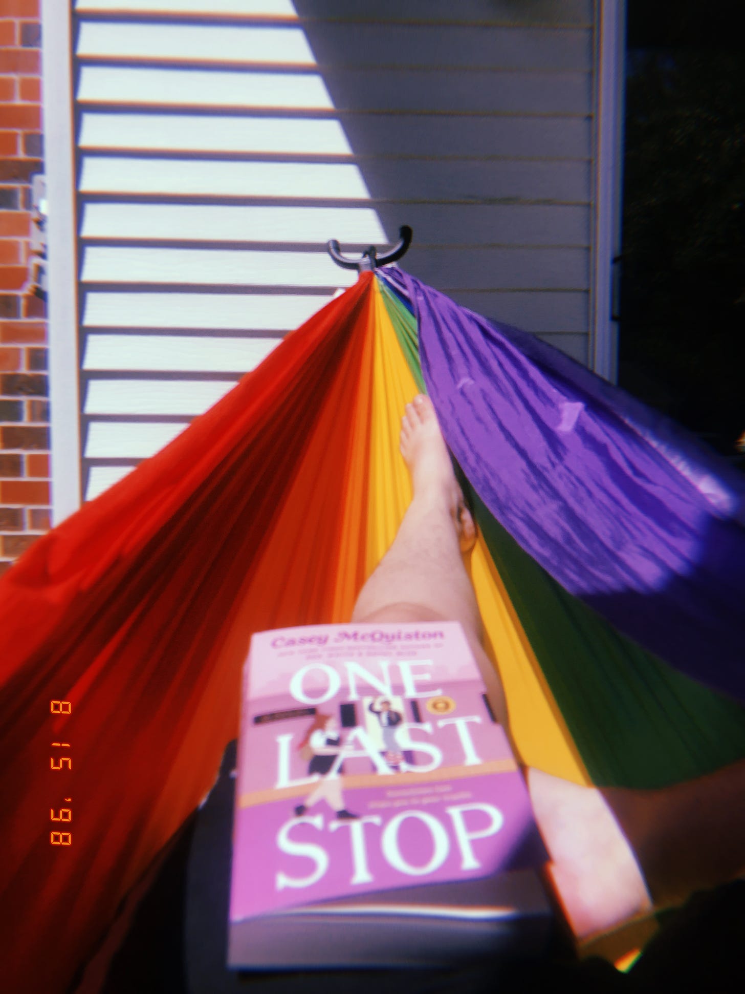 Blurry photo of Emily from the hips down, laying in a rainbow hammock. One Last Stop rests on her thigh.