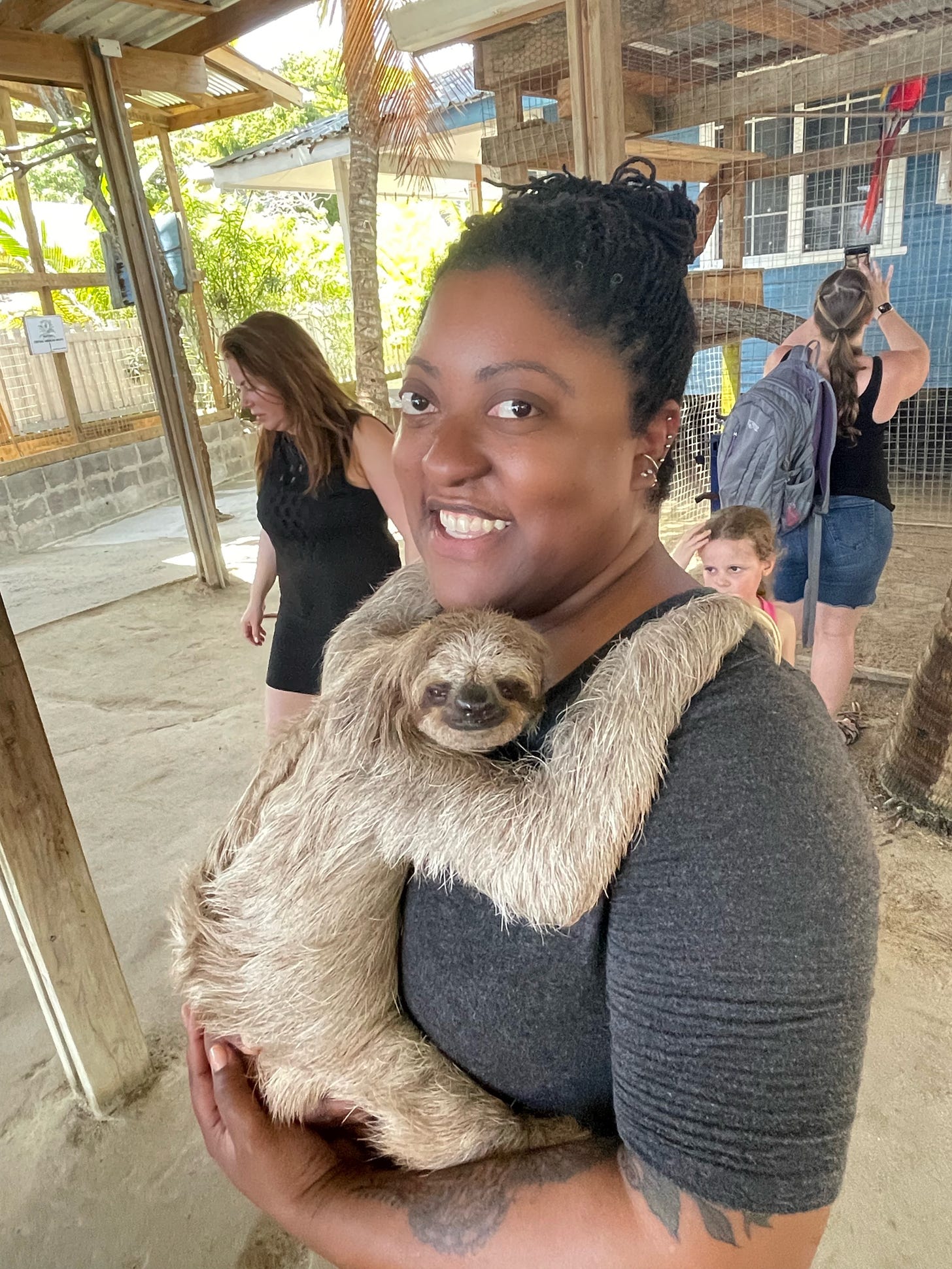 At an animal sanctuary a three toed sloth is held by a smiling Black person with microdreads in a bun