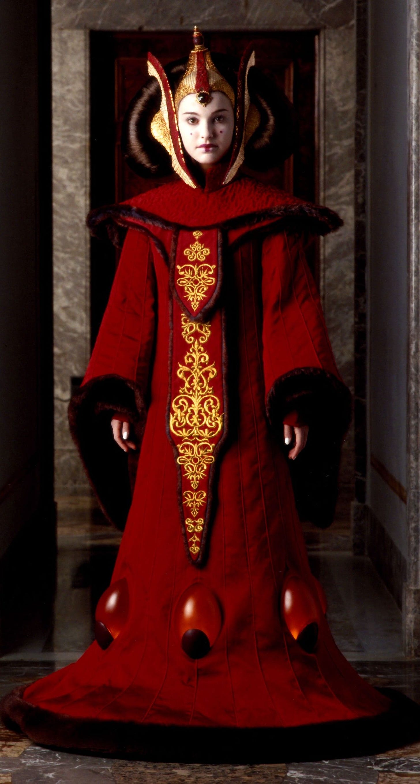 Imagem de Padmé Amidala. Uma mulher jovem e branca, usando um vestido vermelho que cobre do pescoço até seus pés. O vestido é largo e arredondado, dando à ela um formato meio cônico, com ombreiras largas e mangas largas e longas. No centro, há um detalhe em dourado que cobre quase todo o vestido. O rosto dela está pintado de branco, com alguns detalhes em vermelho, como a boca e uma pintinha em cada bochecha. Na cabeça, ela usa um tipo de chapéu/coroa dourado com detalhes em vermelho. A peça cobre todo seu crânio. Na parte central, ele se eleva da base larga e se afina no topo. Nas laterais, dois longos pedaços que vão do seu pescoço até acima de sua cabeça.