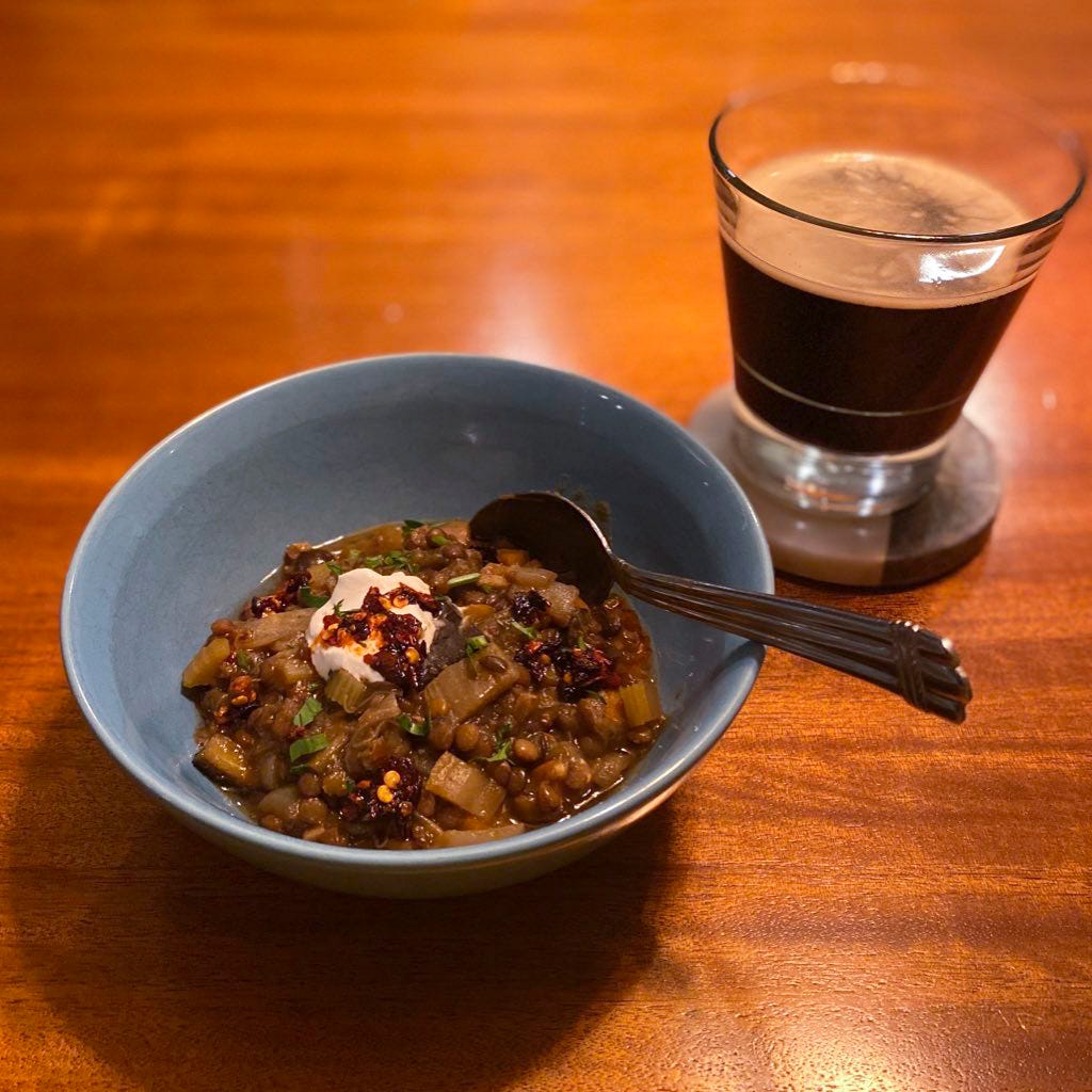 A blue bowl of lentil & eggplant stew with sour cream and chili crisp on top. A glass of dark beer rests on a coaster to its right.