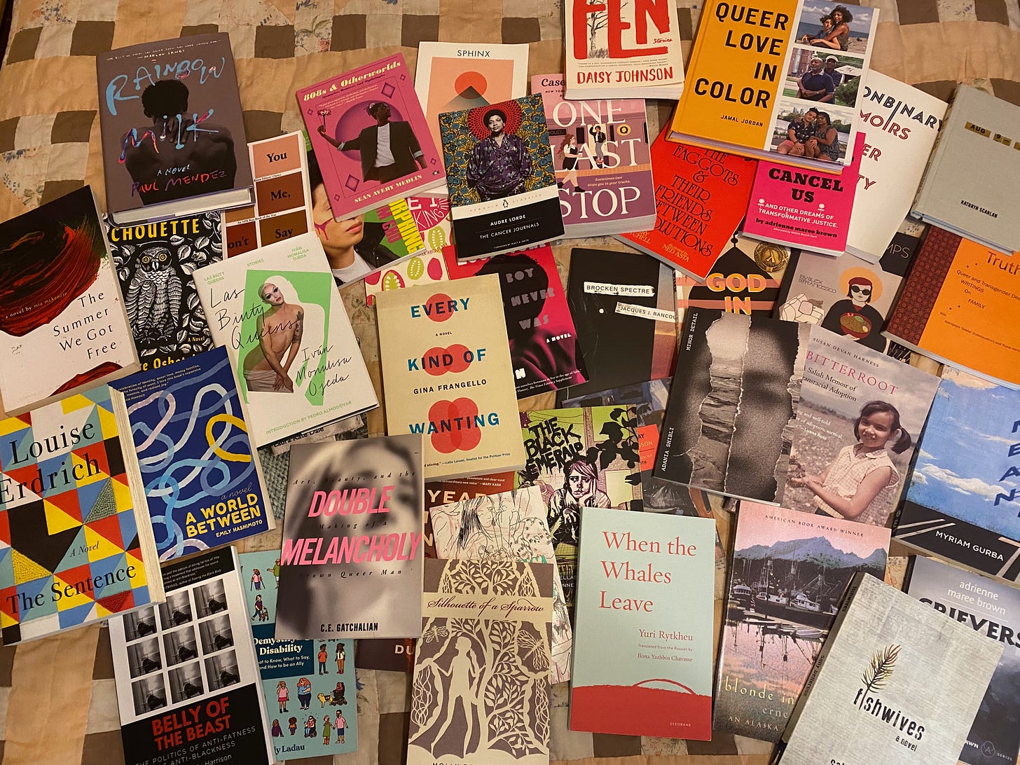 A huge pile of books scattered across a brown and pink quilt. A few of titles: Rainbow Milk, One Last Stop, Queer Love inColor, Double Melancholy, The Sentence, When the Whales Leave, and The Summer We Got Free.