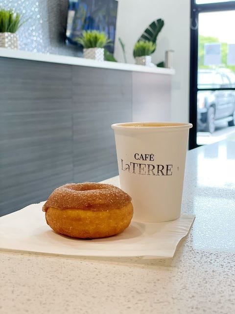 An old-fashioned donut with a chocolate frosting sits on a napkin in front of a white takeout coffee mug stamped "Cafe LaTerre." They both sit on a white marble countertop with dark faux woodstained cabinets in the background.