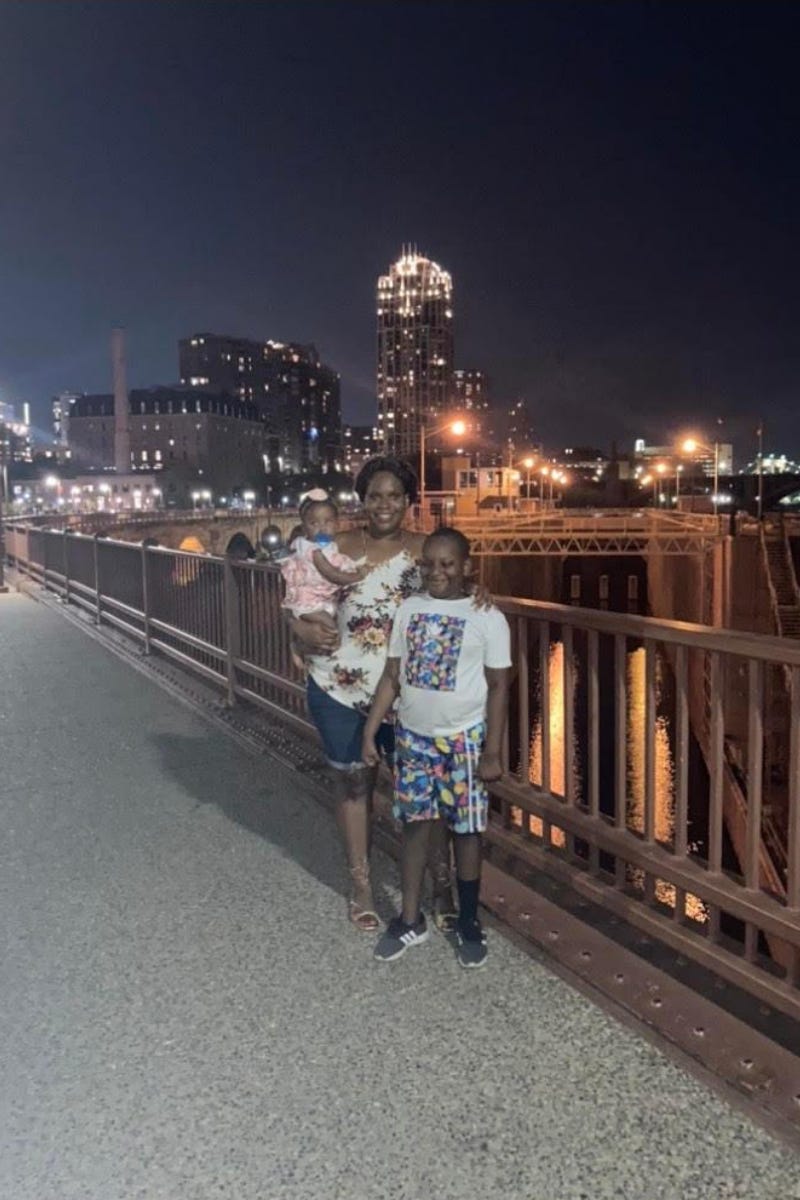 A woman standing on a bridge, holding a young child and standing next an older child with her hand on his sholder, with night sky and city lights in the background.