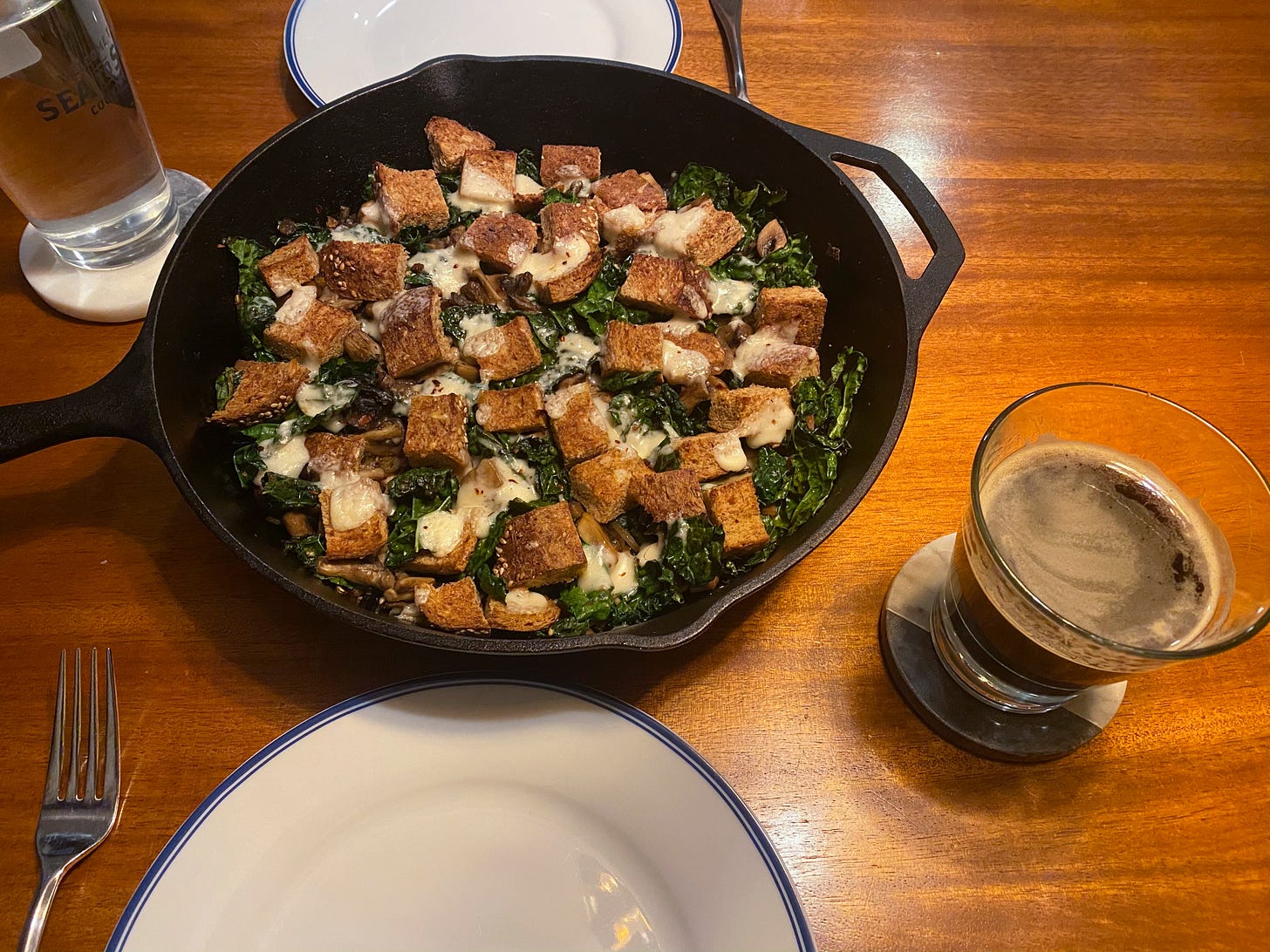 A large cast-iron pan full of kale and mushrooms with brown squares of toast and melted cheese on top. On either side of the pan is a white plate with a blue rim, and next to the plate in the foreground is a half-full glass of dark beer on top of a coaster.