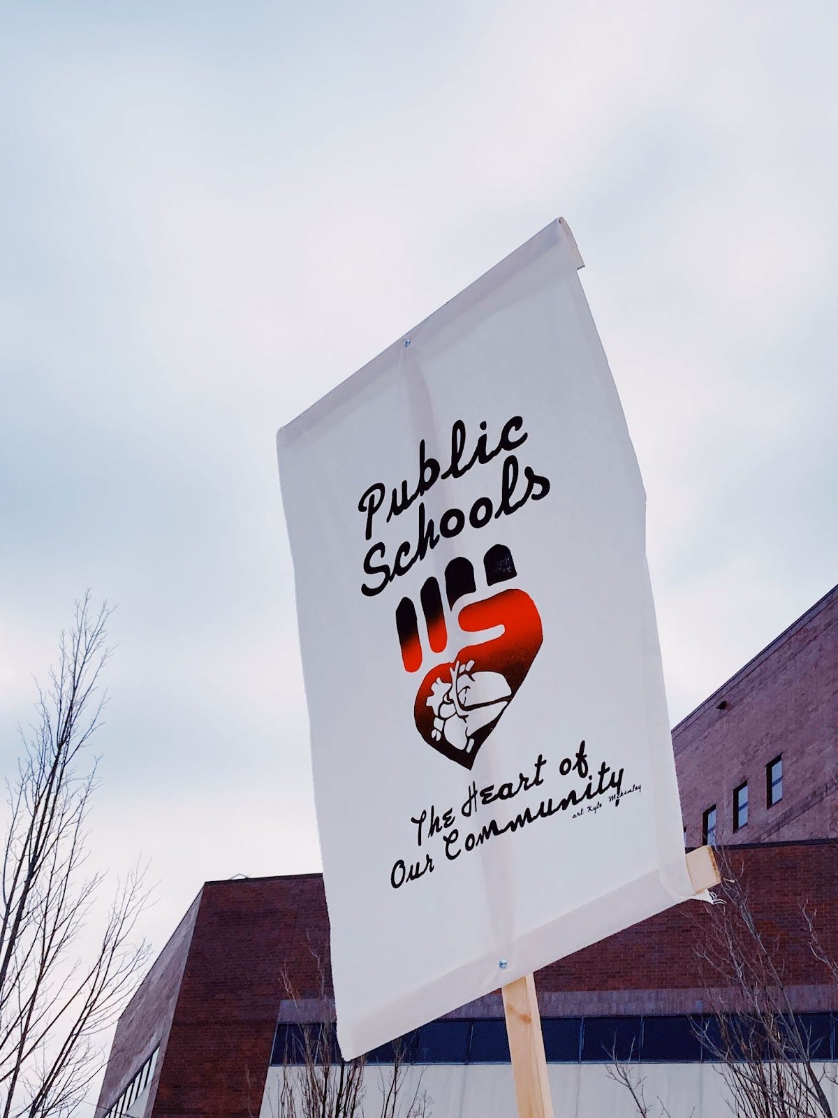 "Public Schools, the heart of our community" is written on black script on a white tarp sign with a logo of a red and black fist with an anatomical heart in the palm.