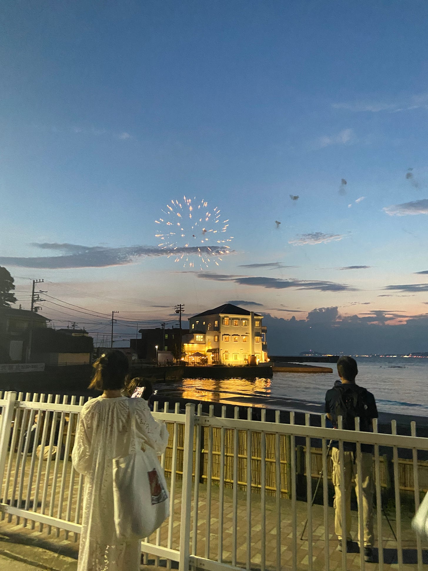 sunset fireworks over the ocean, a white house on the coastline as two people stand in the foreground taking pictures