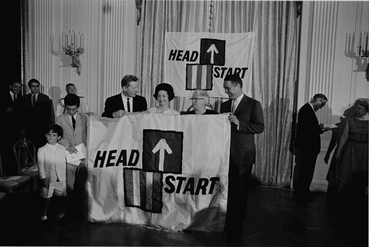 Four white people pose while holding up a banner that reads "Head Start" with an arrow pointing upward