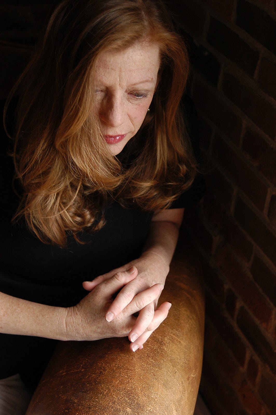 Susan Orlean portrait, her eyes cast down, face partly obscured by her hair, hands folded on chair arm.