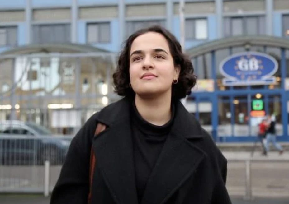 A person with very light skin stands in a black coat outside in front of shops on a street during the daytime. She is looking into the distance behind the camera, slightly smiling. She has dark chin length curly hair and dark eyes. She is wearing gold earrings. She has a brown shoulder strap on her shoulder. Behind her on the left is a dark car. Behind her on the right are two people walking past.