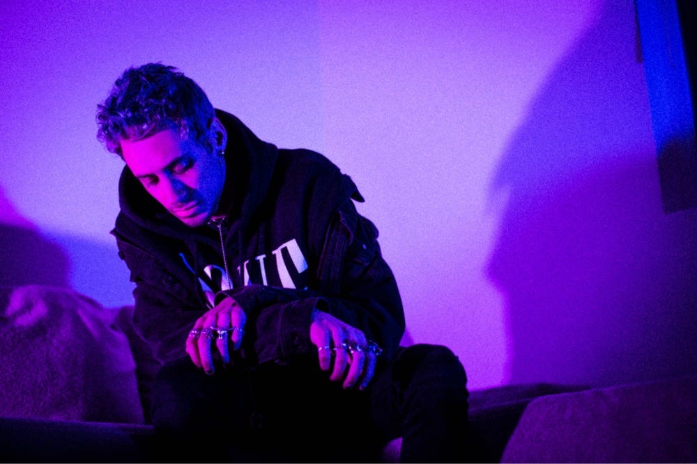 cøzybøy posing with his hands crossed and face looking down. The photo is shrouded in darkness with pink and blue tones.