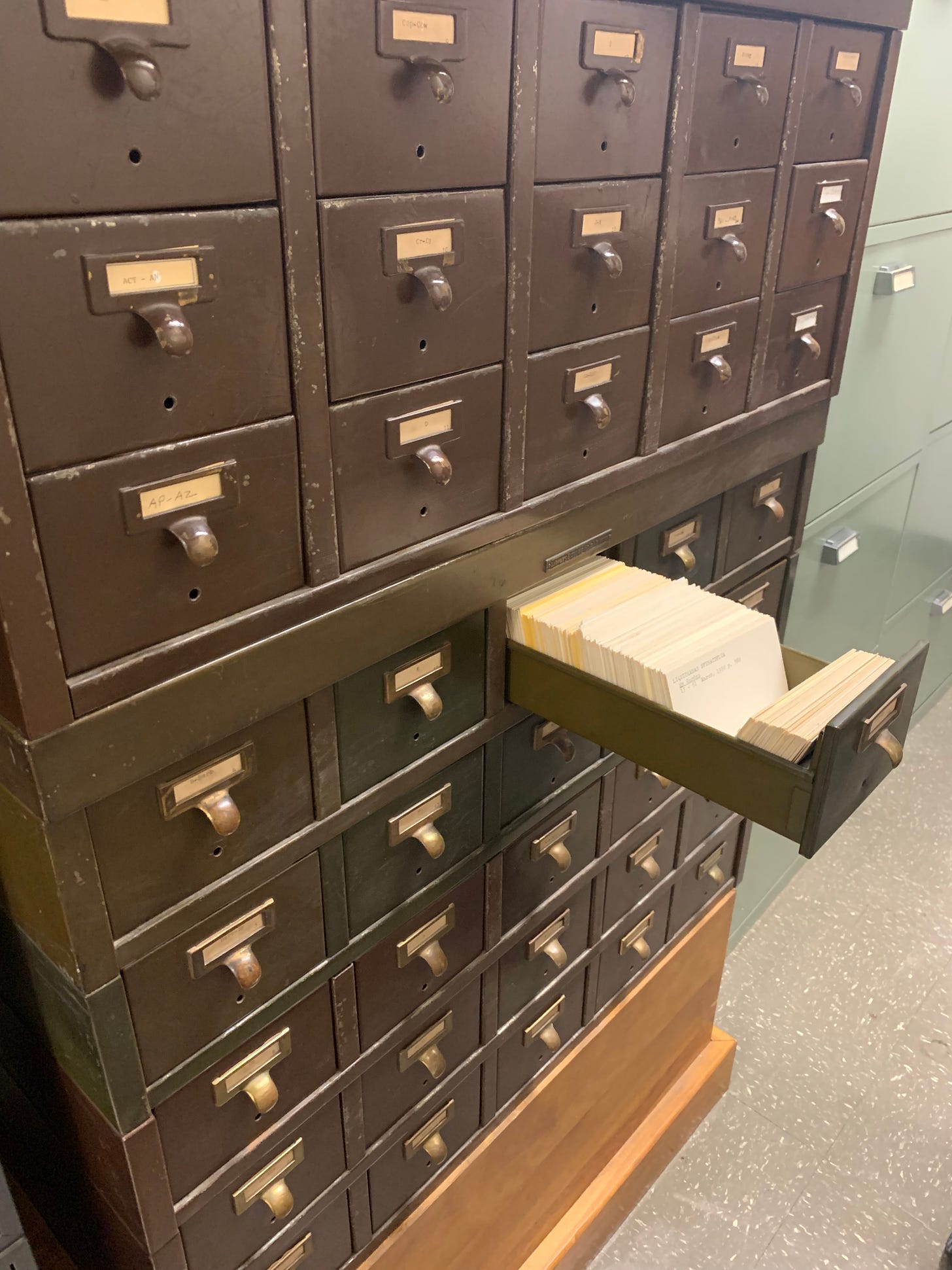 A brown cabinet with yellowed labels on the front of each drawer. A drawer is pulled open and is filled with index cards.