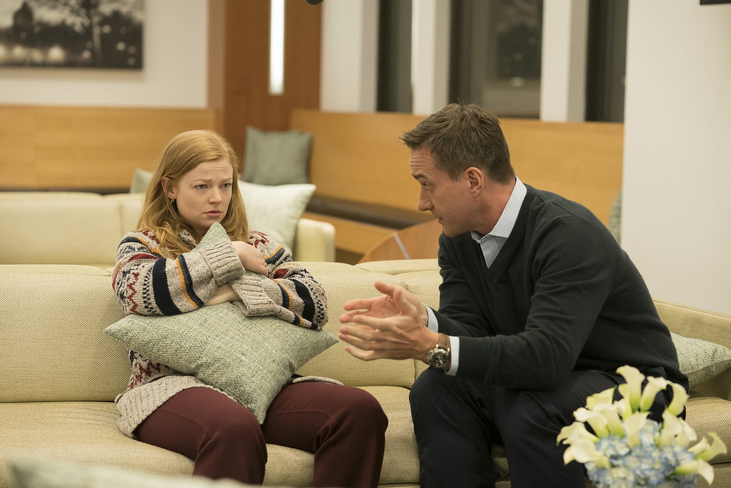 Sarah Snook as Shiv Roy sitting and holding a pillow (left) and Matthew Macfadyen as Tom Wambsgans talking to Shiv and sitting on her left (right) in SUCCESSION.