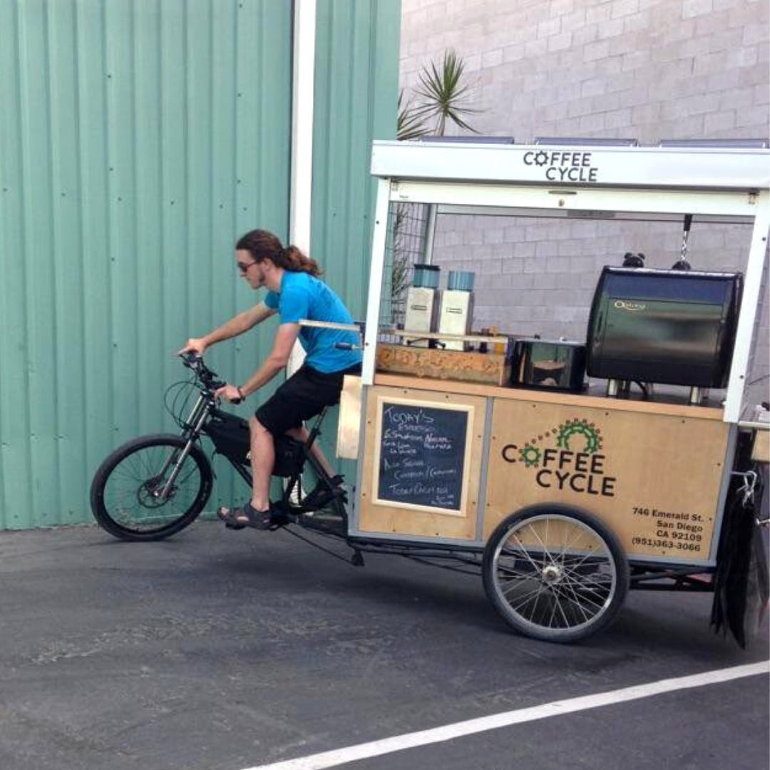 A full coffee cart complete with espresso machine being pulled behind a bicycle by a white man with a ponytail wearing a blue tee shirt and black shorts.