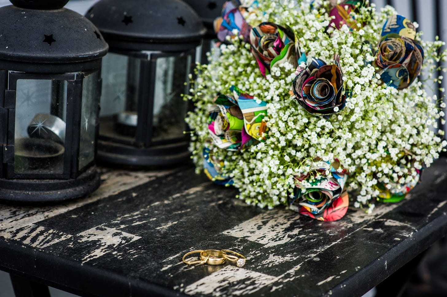 Close up photo of three gold wedding rings sitting on a distressed table, with a bouquet of rolled comic book flowers surrounded by baby's breath. 