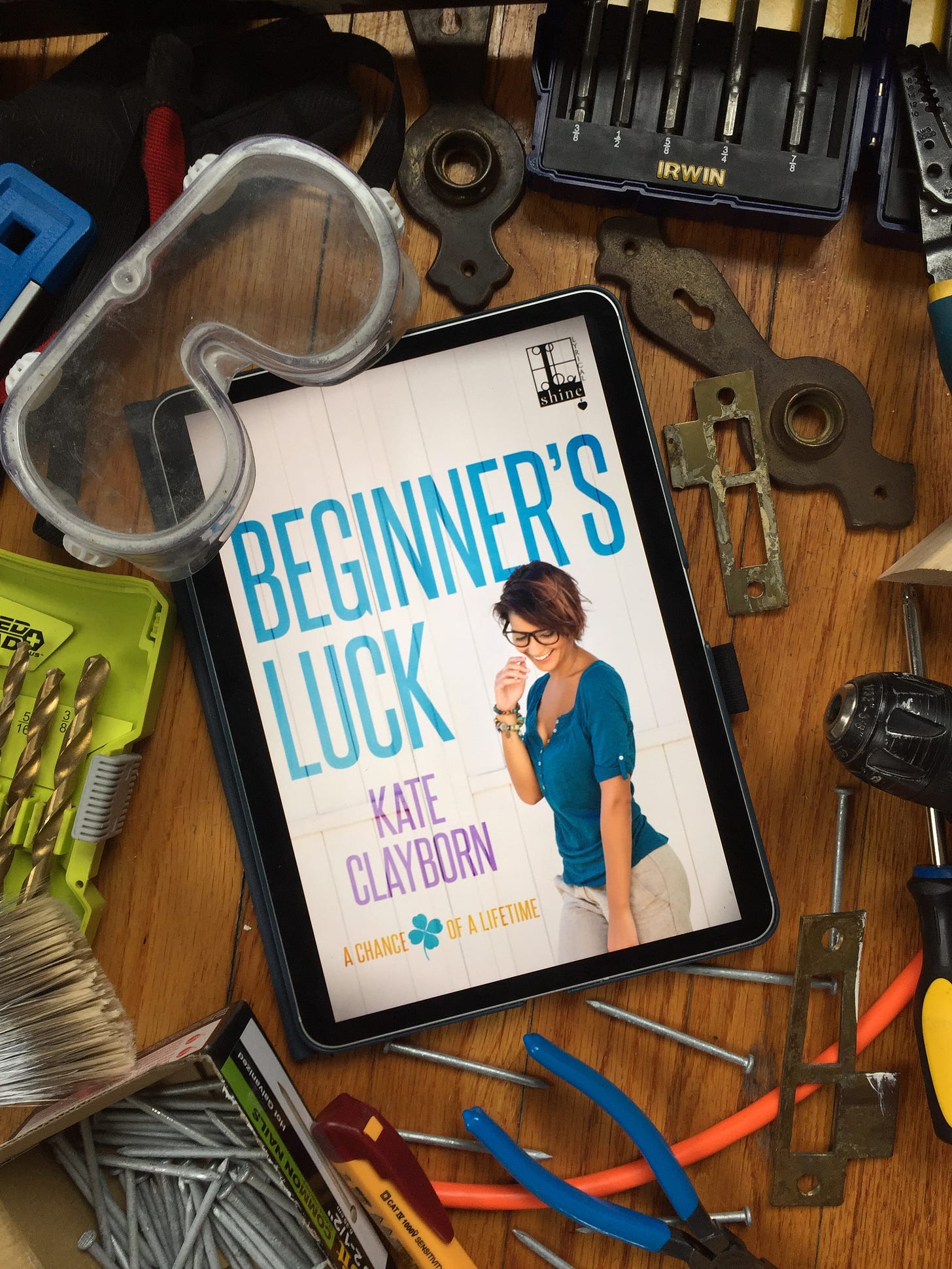 An ereader displaying the cover of Kate Clayborn's Beginner's Luck, surrounded by nails, tools, safety goggles, and door findings on a wood floor.