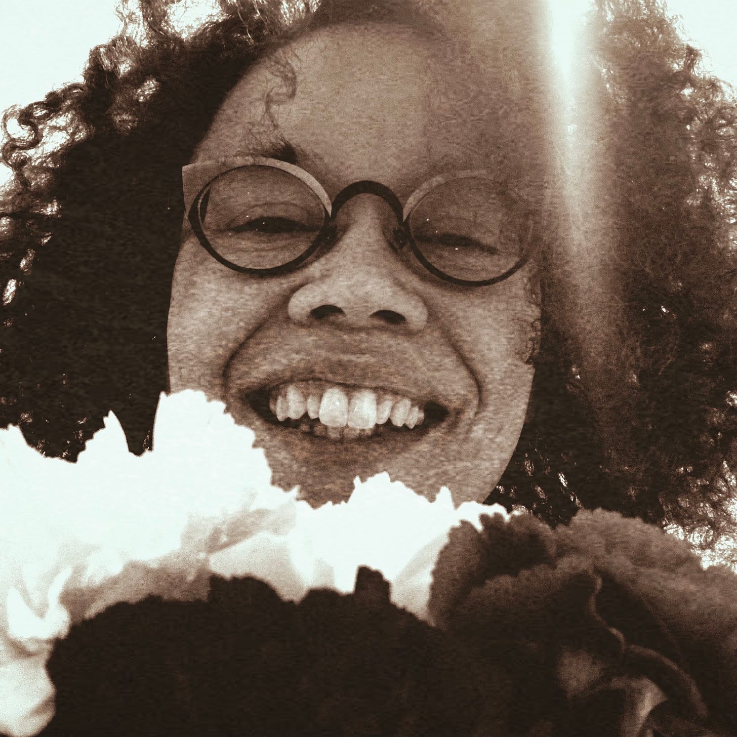 Close up of Ianna's face in sepia, smiling widely and with teeth. Dark curly hair, two-tone glasses frames. Three big flowers in the foreground, just below Ianna's mouth.