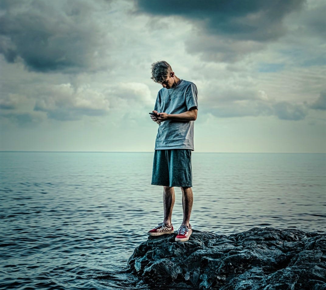 moody photo with stormy sky young man wearing t-shirt and shorts standing on rocks with ocean behind while he looks at his phone moody photo with stormy sky young man wearing t-shirt and shorts standing on rocks with ocean behind while he looks at his phone