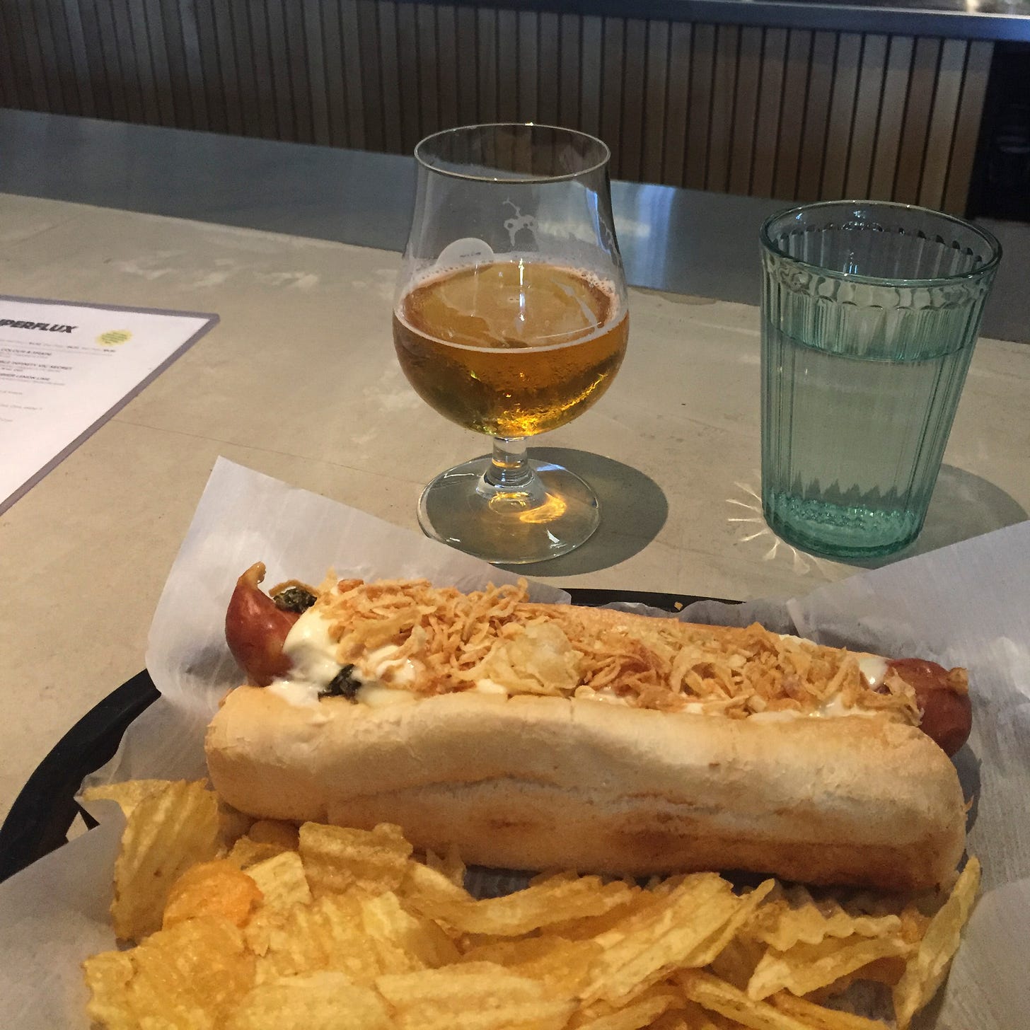 In a black plastic basket lined with parchment, a small pile of ripple chips is in the foreground next to a hot dog, which is covered with crispy onions, mayo, and chimichurri. A tulip glass of beer is on the bar behind it, next to a glass of water.