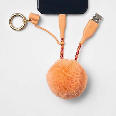 Product photo from the Target website of a pom pom keychain charger. It's coral colored with a big fluffy ball on the bottom and an orange and pink braided cord with one end plugged into a phone and the other end, a USB connector, free. There's a round keychain on it as well.